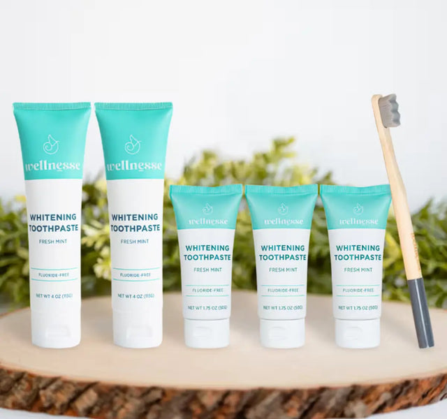 Whitening toothpaste fluoride-free by Wellnesse in five fresh mint tubes of two sizes arranged on a wood slice next to a bamboo charcoal toothbrush.