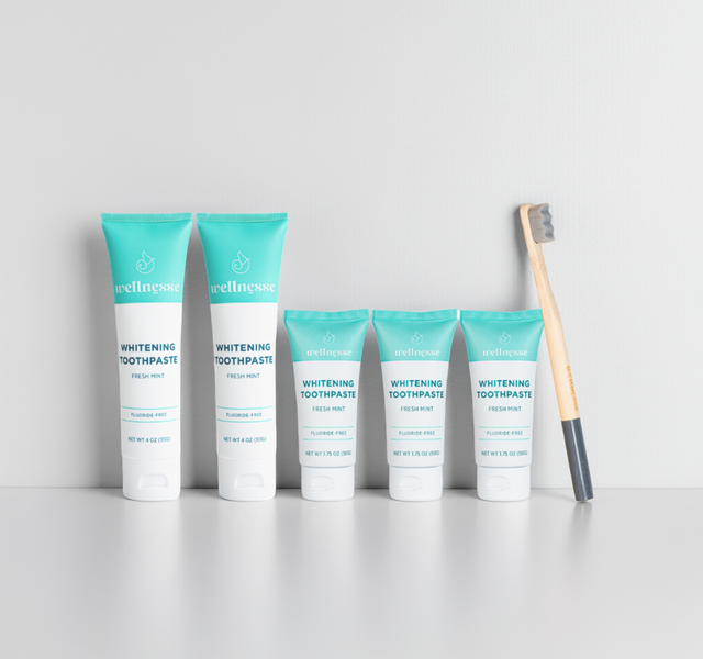Fluoride-free whitening toothpaste by Wellnesse in five fresh mint tubes of two sizes lined up beside a bamboo charcoal toothbrush on a white surface.