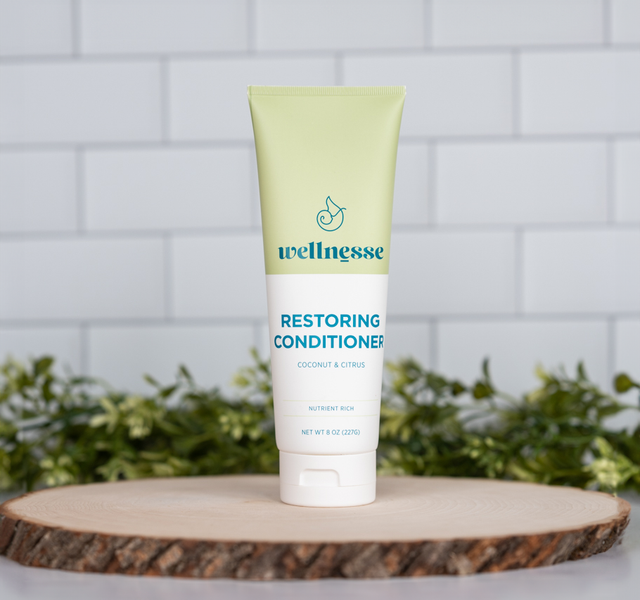 Wellnesse restoring conditioner in coconut citrus scent on a wood slice with white tile and green plants behind