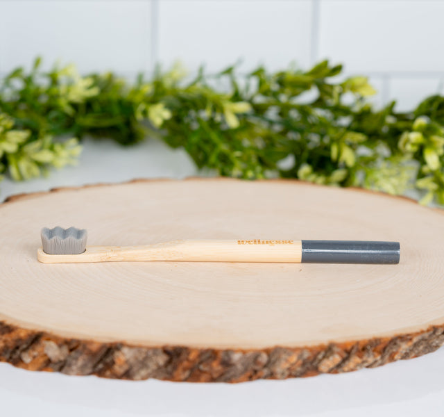 Bamboo charcoal toothbrush by Wellnesse with gray bristles lying flat on a wood slice with green plants in the background.