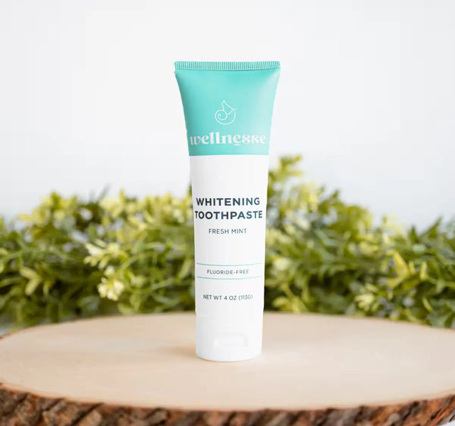 Wellnesse natural whitening toothpaste in fresh mint with teal cap on a wood slice with greenery behind