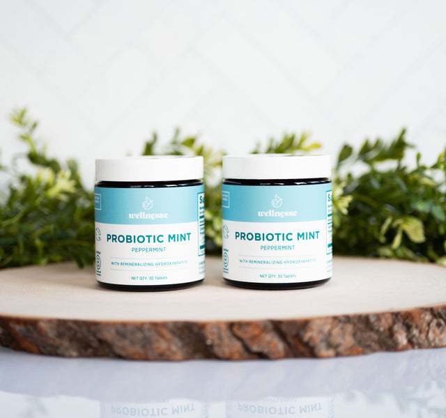 Two jars of Wellnesse peppermint probiotic mints with hydroxyapatite on a wood slice with greenery.