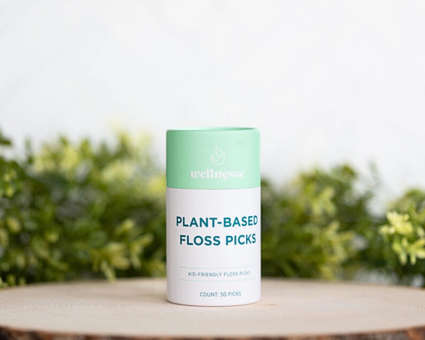 Floss picks in a mint green and white Wellnesse container labeled plant-based, displayed on a wooden surface with greenery.