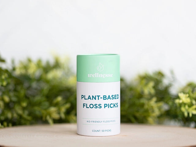 Floss picks in a mint green and white Wellnesse container labeled plant-based, displayed on a wooden surface with greenery.