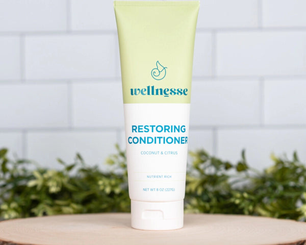 Restoring conditioner with coconut and citrus by Wellnesse, standing on a wood slice with greenery and tiled background.