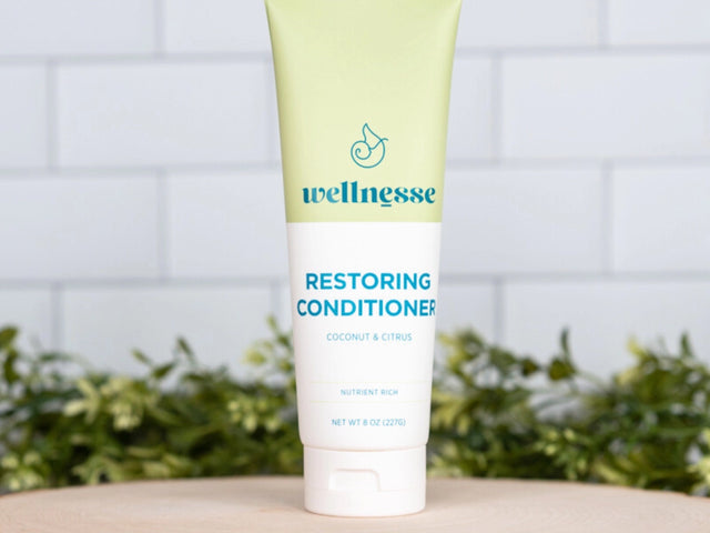 Restoring conditioner with coconut and citrus by Wellnesse, standing on a wood slice with greenery and tiled background.