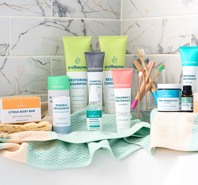 Wellnesse natural personal care product collection including shampoo, charcoal toothpaste, children's toothpaste, oral probiotics, mineral deodorant, and body bar arranged on a bathroom counter with teal towels.