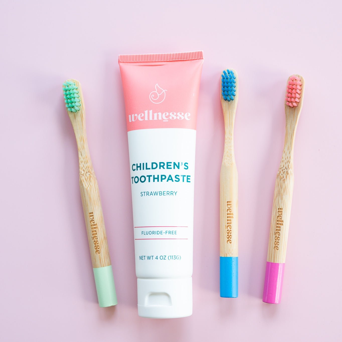 Strawberry natural ingredient toothpaste for kids by Wellnesse, displayed with colorful bamboo toothbrushes on a pink background
