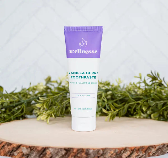 Wellnesse vanilla toothpaste in berry flavor standing on a wood slice surrounded by fresh greenery