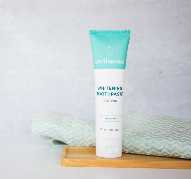 Wellnesse whitening toothpaste standing on a bamboo tray with a folded teal towel from the oral care set