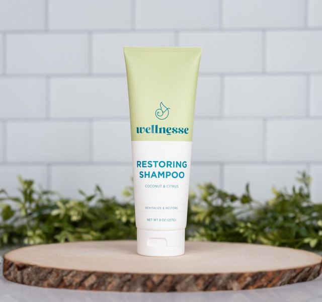 Wellnesse restoring shampoo in coconut citrus scent on a wood slice against a white tile backdrop with green plants
