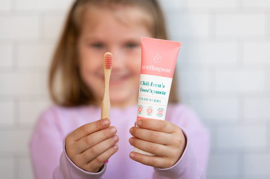 Kids strawberry natural and organic toothpaste held by a smiling child alongside a bamboo toothbrush with pink bristles.